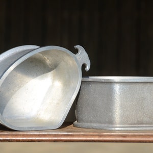 Puede incluir: Dos moldes para hornear de aluminio plateado, uno con forma redondeada y el otro con forma rectangular. Ambos moldes tienen un acabado liso y metálico.