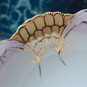 May include: A white porcelain bowl with a pink rim and gold accents. The bowl has a scalloped edge and a decorative design of stylized leaves and a fan-shaped pattern.