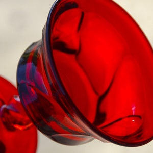 May include: A red glass goblet with a wide, round bowl and a short stem. The goblet is made of clear glass with a red tint.
