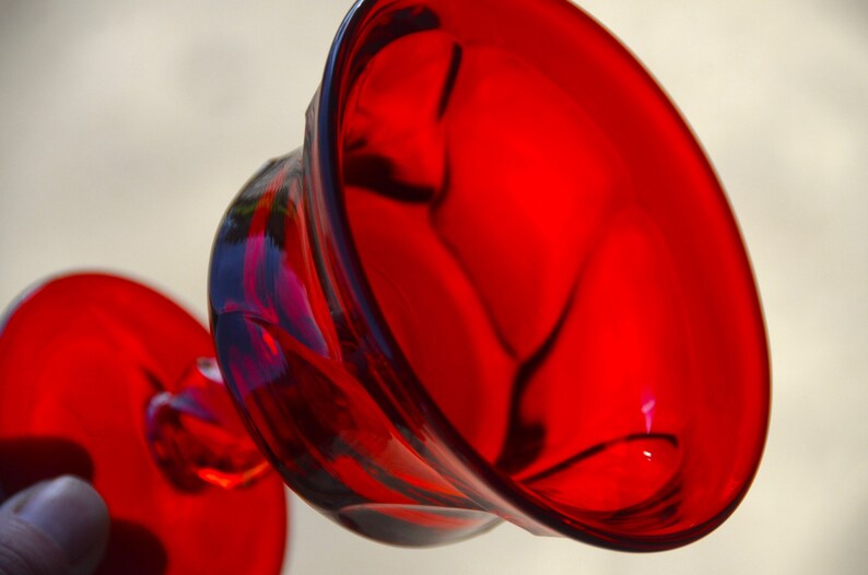 May include: A red glass goblet with a wide, round bowl and a short stem. The goblet is made of clear glass with a red tint.