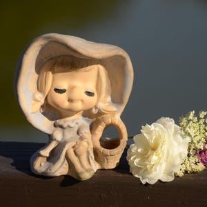 Może przedstawiać: Ceramiczna figurka dziewczynki w czapeczce, trzymającej mały koszyk. Figurka jest pomalowana w delikatnych pastelowych kolorach i ma słodki, niewinny wyraz twarzy. Figurka stoi na drewnianej powierzchni z białą różą i zielonym listowiem.