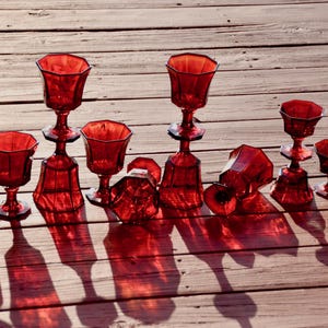Pode incluir: Uma coleção de cálices e copos de haste em vidro vermelho translúcido com um design facetado octogonal. Alguns copos estão na vertical enquanto outros estão posicionados de lado, exibindo o padrão geométrico do vidro.