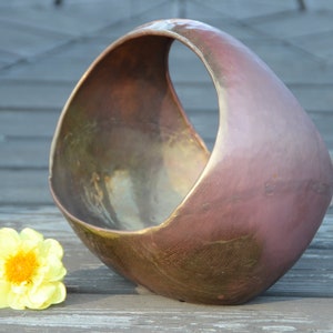 May include: A large, round, copper-colored ceramic planter with a hole in the side. The planter has a rustic, aged look and is sitting on a wooden surface.