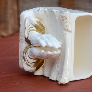 May include: White ceramic figurine of two hands clasped in prayer, with gold accents. The figurine is shaped like a book with a hollow space for holding items.