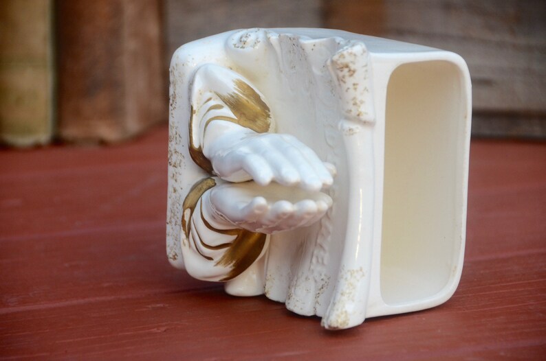 May include: White ceramic figurine of two hands clasped in prayer, with gold accents. The figurine is shaped like a book with a hollow space for holding items.