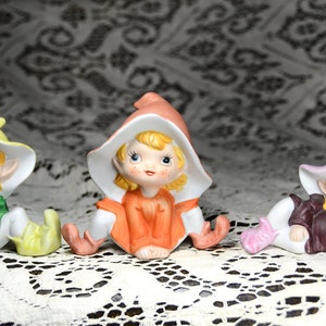May include: Three porcelain figurines of elves with colorful hats. The elf on the left is wearing a yellow hat, the elf in the middle is wearing an orange hat, and the elf on the right is wearing a pink hat.