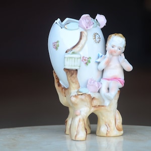 May include: A white porcelain egg-shaped vase with a cherub figurine. The vase is decorated with pink roses and floral accents. The cherub has a pink sash and is perched on the side of the vase. The base is a tree trunk design.