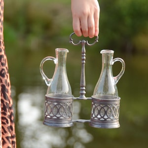 May include: A silver metal bottle holder with two clear glass bottles. The holder has a decorative design and is being held by a hand.