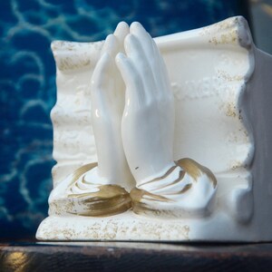 May include: White ceramic figurine of two hands clasped in prayer, with gold accents. The figurine is set against a blue background.