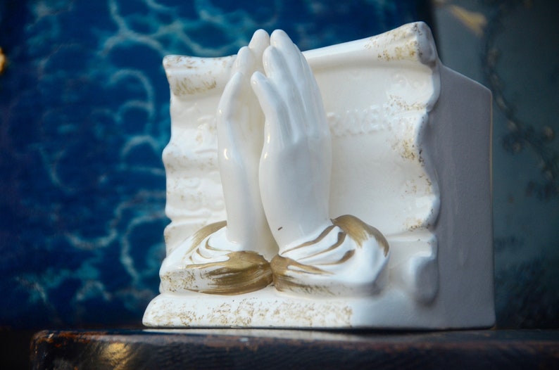 May include: White ceramic figurine of two hands clasped in prayer, with gold accents. The figurine is set against a blue background.