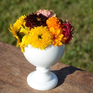 Puede incluir: Un jarrón de cerámica blanco con un cuenco redondo y una base de pedestal, lleno de un ramo de flores amarillas, naranjas, rojas y marrones.