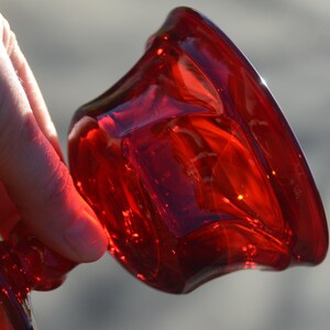 May include: A deep red glass candle holder with a wide, flared base and a rounded top. The holder is made of thick, clear glass with a textured surface.