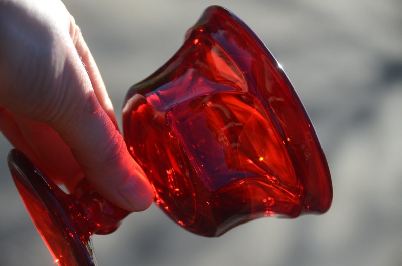 May include: A deep red glass candle holder with a wide, flared base and a rounded top. The holder is made of thick, clear glass with a textured surface.