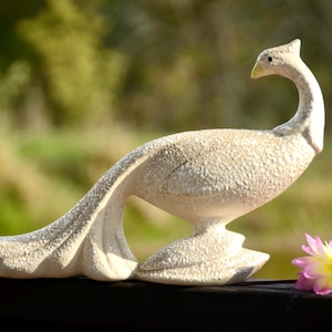 Op de afbeelding: Een wit keramiek pauw beeldje met een getextureerd oppervlak. De pauw staat op een houten oppervlak met een roze bloem op de voorgrond.