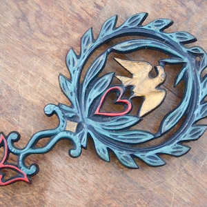 Könnte beinhalten: Ein gusseiserner Topf-Untersetzer mit blauer, grüner und goldener Oberfläche. Der Untersetzer ist in Form eines Kranzes mit einem Vogel und einem Herzen in der Mitte.