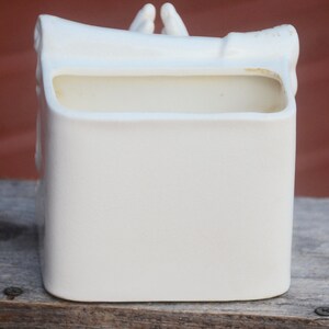 May include: White ceramic planter shaped like a pair of hands holding a scroll. The planter has a rectangular opening for planting.