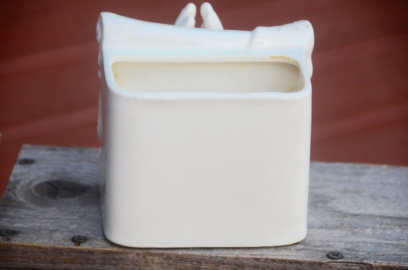 May include: White ceramic planter shaped like a pair of hands holding a scroll. The planter has a rectangular opening for planting.