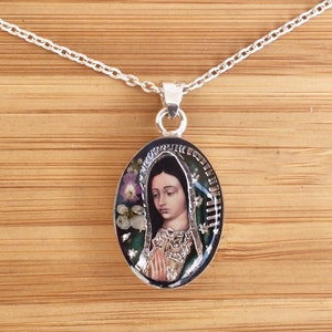 Colgante ovalado de la Virgen de Guadalupe: verde con detalles en plata, flores reales en miniatura moradas y blancas engastadas en resina con base de plata esterlina