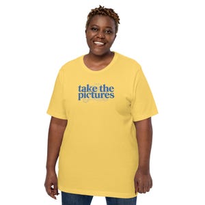 May include: A short-sleeved yellow t-shirt featuring the blue text "take the pictures" printed on the center of the chest. The design includes a faint, light-colored line drawing of a vintage camera positioned directly behind the blue lettering.