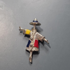 May include: A silver-toned brooch in the shape of a stylized scarecrow. The scarecrow has a hat, arms outstretched, and patches of yellow, blue, and red on its coat. The brooch is detailed with textured metal and small embellishments.