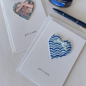 May include: Two blank greeting cards with fabric hearts attached. The hearts are decorated with different patterns and have a ribbon tied around them. The text "just a note" is written on each card.