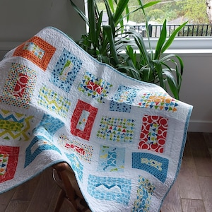 May include: A patchwork quilt with a white base and vibrant square patterns in orange, blue, red, and green. The quilt is draped over a wooden chair, highlighting its detailed design and soft texture. The quilt's dimensions are approximately 152 x 178 cm.
