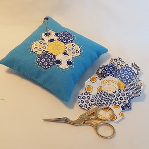 May include: A blue fabric pincushion with a yellow and blue flower design. The pincushion is filled with pins and is sitting on a white surface. There are also several yellow and blue fabric pieces on the surface, along with a pair of gold scissors.