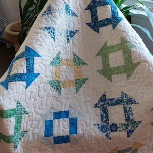 May include: A handmade quilt with a white background and colourful geometric patterns. The quilt features squares and arrow-like shapes in shades of blue, green, and yellow. The quilt is draped over a chair.