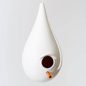 Może przedstawiać: Biały ceramiczny domek dla ptaków w kształcie łzy z okrągłym otworem wejściowym i pomarańczowym drążkiem.