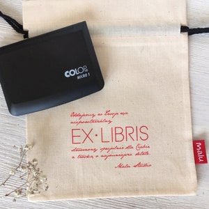 Könnte beinhalten: Eine weiße Stofftasche mit einem schwarzen Kordelzug. Die Tasche hat einen roten Stempel mit der Aufschrift "EX LIBRIS" und "Malu Studio" in Kursivschrift. Die Tasche enthält außerdem ein schwarzes Stempelkissen mit der Aufschrift "COLOP MICRO 1".
