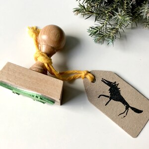 Op de afbeelding: Houten stempel met een groen boomontwerp en een gele string bevestigd aan een houten handvat. De stempel wordt gebruikt om een ​​zwart-witte wolfillustratie op een bruin papieren label te maken.