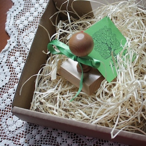 Puede incluir: Un sello de madera con una cinta verde y un dise&ntilde;o de &aacute;rbol en una tarjeta verde. El sello est&aacute; en una caja marr&oacute;n con papel triturado.