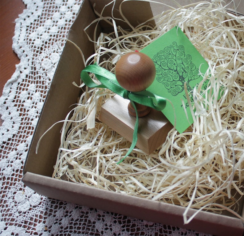 Puede incluir: Un sello de madera con una cinta verde y un dise&ntilde;o de &aacute;rbol en una tarjeta verde. El sello est&aacute; en una caja marr&oacute;n con papel triturado.