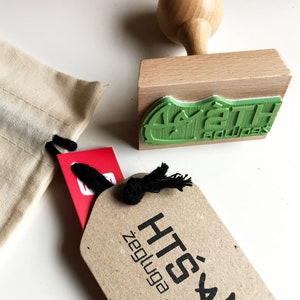 Op de afbeelding: Een bruin papieren label met zwarte tekst die "HTS'" en "ZeugYoga" luidt. Een rood label met zwarte tekst is zichtbaar op de achtergrond. Een houten stempel met een groene rubberen stempel die "Spath Boulder" luidt, is ook zichtbaar op de achtergrond.