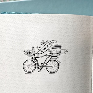 May include: Black and white illustration of a bicycle with a basket of flowers and a stack of books tied to the back. The bicycle has a banner that reads "Ex Libris Bernadette Flamanti".