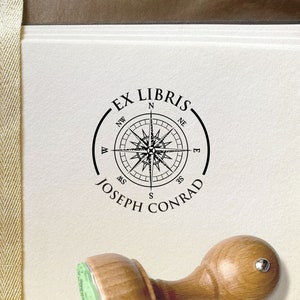 Wind Rose Personalized Ex Libris Stamp. Round wooden stamp with the knob on top of the wooden handle. Vintage-style compass illustration with cardinal directions. On top of the illustration is EX LIBRIS and below is the name of the owner of the stamp