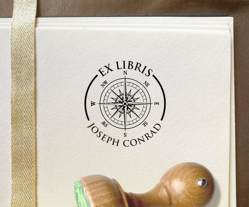 Wind Rose Personalized Ex Libris Stamp. Round wooden stamp with the knob on top of the wooden handle. Vintage-style compass illustration with cardinal directions. On top of the illustration is EX LIBRIS and below is the name of the owner of the stamp