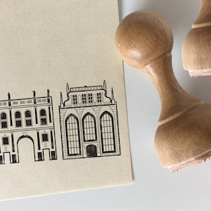 Può includere: Due timbri in gomma di legno con disegni di edifici in inchiostro nero. I timbri sono su uno sfondo beige. I disegni sono di un edificio con un grande arco e un edificio con una grande finestra.