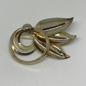 May include: A gold-toned brooch with a circular design and three leaf-shaped accents. The brooch has a pin on the back for attaching to clothing.