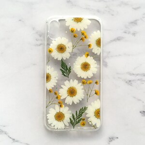 May include: A clear phone case with a pressed flower design. The case features white daisies and yellow centers, pressed into the clear plastic.