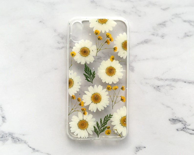 May include: A clear phone case with a pressed flower design. The case features white daisies and yellow centers, pressed into the clear plastic.