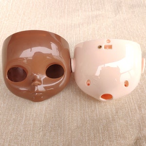 Puede incluir: Dos cabezas de mu&ntilde;eca, una en un tono marr&oacute;n oscuro y la otra en rosa claro. Ambas tienen grandes cuencas de los ojos, una nariz y una boca. La cabeza marr&oacute;n tiene un acabado brillante, mientras que la rosa tiene un acabado mate. Probablemente sean componentes para una mu&ntilde;eca.