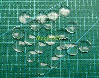 May include: A collection of clear round glass cabochons, scattered on a green cutting mat. The cabochons are various sizes and are perfect for crafting projects.