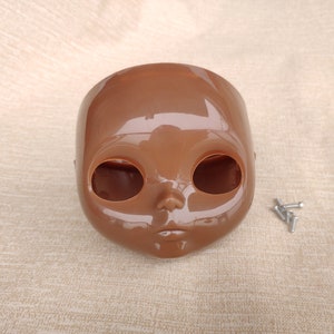 Puede incluir: Una cabeza de mu&ntilde;eca de pl&aacute;stico marr&oacute;n con grandes cuencas de los ojos y una peque&ntilde;a nariz y boca. Varios tornillos plateados peque&ntilde;os est&aacute;n a la derecha de la cabeza de la mu&ntilde;eca. La cabeza de la mu&ntilde;eca es de color s&oacute;lido y tiene un acabado brillante.