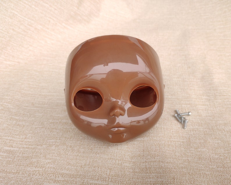Puede incluir: Una cabeza de mu&ntilde;eca de pl&aacute;stico marr&oacute;n con grandes cuencas de los ojos y una peque&ntilde;a nariz y boca. Varios tornillos plateados peque&ntilde;os est&aacute;n a la derecha de la cabeza de la mu&ntilde;eca. La cabeza de la mu&ntilde;eca es de color s&oacute;lido y tiene un acabado brillante.