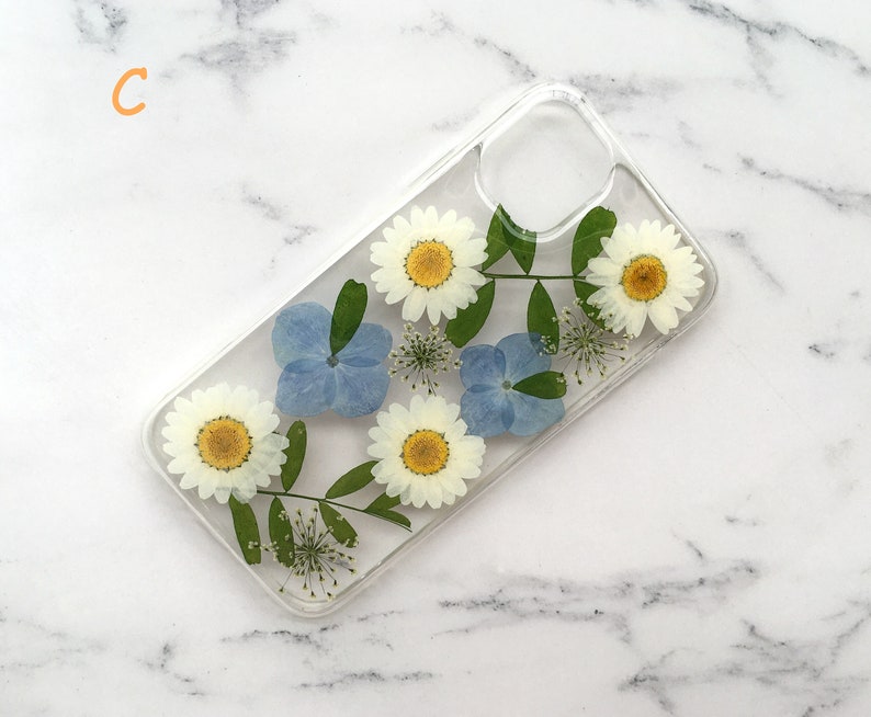 May include: A clear phone case with pressed flowers. The case features white daisies and blue hydrangeas.