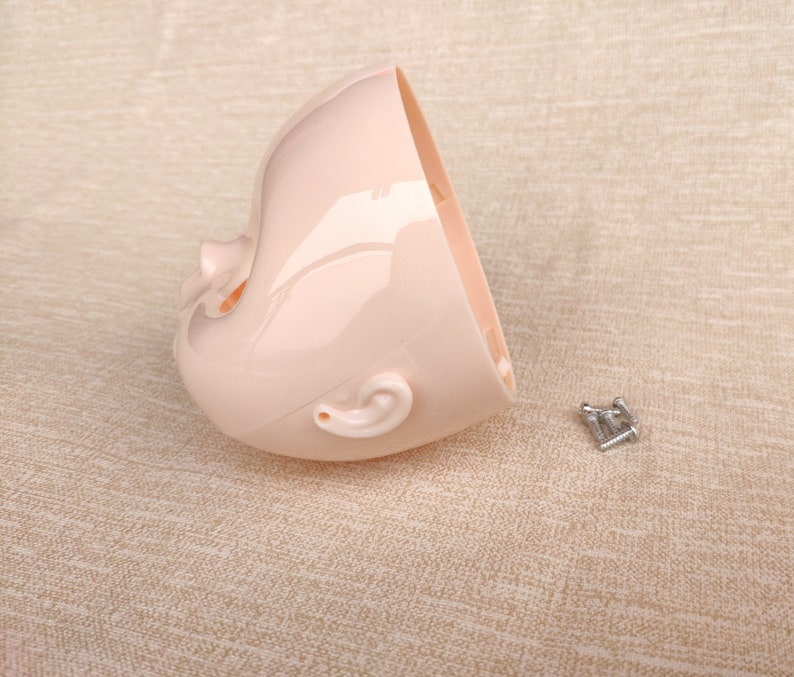 Puede incluir: Una cabeza de mu&ntilde;eca de pl&aacute;stico rosa claro, cortada por la mitad, con rasgos faciales visibles, incluyendo una oreja, nariz y cuenca del ojo. Varios tornillos plateados peque&ntilde;os est&aacute;n junto a la cabeza de la mu&ntilde;eca. El fondo es una tela de color beige claro.
