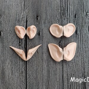 May include: Six pairs of elf ears made of a light pink flexible material. The ears are arranged in three rows on a dark wood background. The ears are for cosplay or costume making.