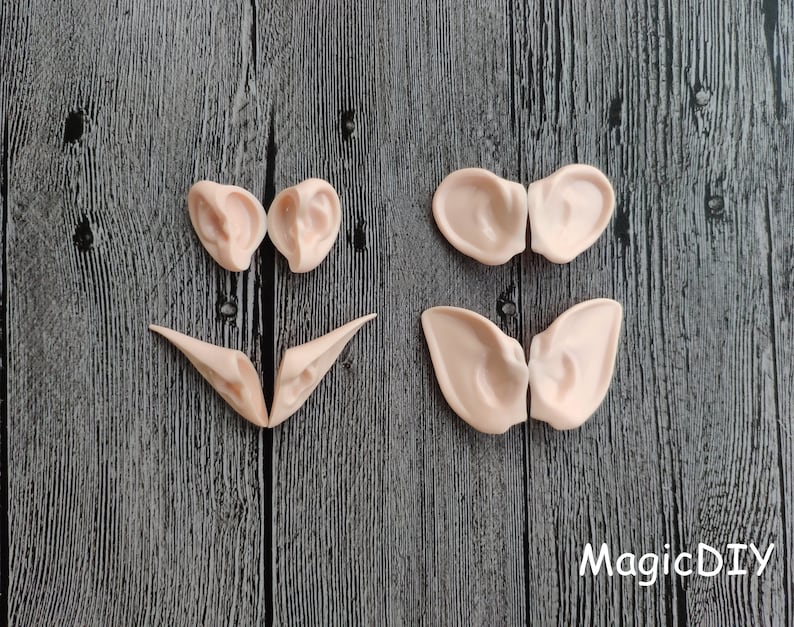 May include: Six pairs of elf ears made of a light pink flexible material. The ears are arranged in three rows on a dark wood background. The ears are for cosplay or costume making.