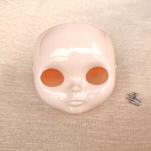 Puede incluir: Una cabeza de mu&ntilde;eca de color rosa p&aacute;lido con grandes cuencas oculares y una boca peque&ntilde;a y cerrada. La cabeza es lisa y brillante, con unos peque&ntilde;os tornillos a la derecha. La cabeza de la mu&ntilde;eca es probablemente para un proyecto de mu&ntilde;eca personalizado.